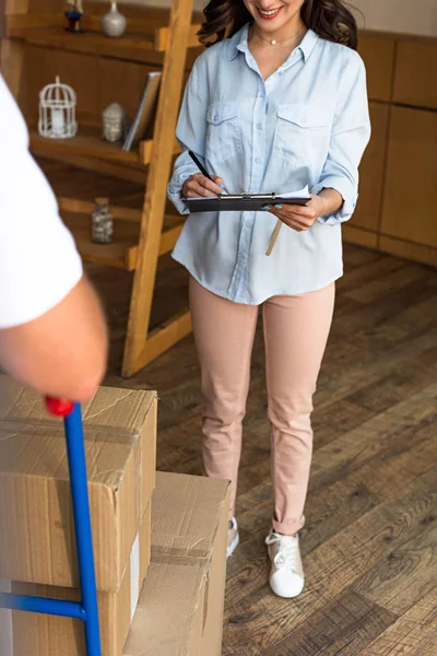 Vue recadrée de femme heureuse papier de signature tout en tenant presse-papiers près du livreur — Photo de stock