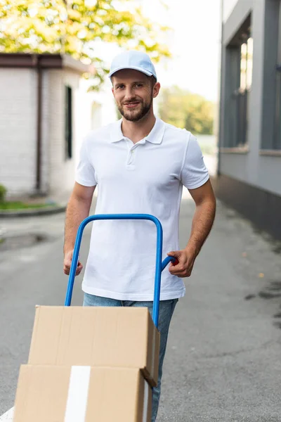 Positive delivery man standing with delivery cart near building — Stock Photo