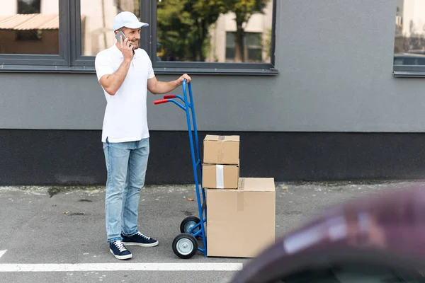 Selektiver Fokus eines glücklichen bärtigen Zustellers, der mit dem Smartphone spricht, während er den Lieferwagen hält — Stockfoto