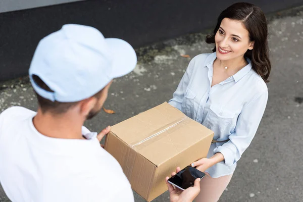 Vista aérea del hombre de entrega en la caja de la tapa que da a la mujer feliz mientras que toma el teléfono inteligente con la pantalla en blanco fuera — Stock Photo