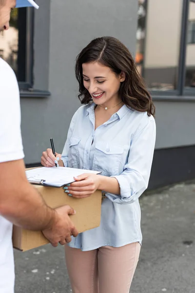 Recortado vista de repartidor hombre celebración caja cerca feliz chica firma papel en portapapeles fuera - foto de stock