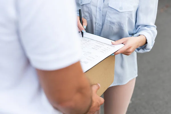 Vue recadrée de livraison homme tenant boîte près fille tenant stylo près presse-papiers à l'extérieur — Photo de stock