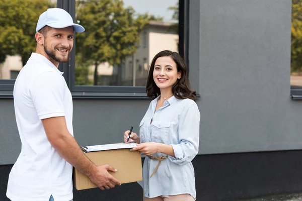 Smiling delivery man holding box near woman signing paper on clipboard outside — Stock Photo