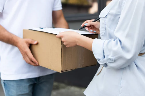 Cropped view of delivery man holding box near woman signing paper on clipboard outside — Stock Photo