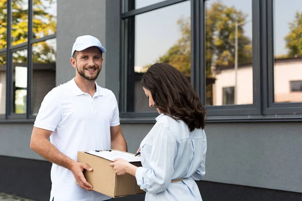 Happy bearded delivery man holding box near woman signing paper on clipboard outside — Stock Photo