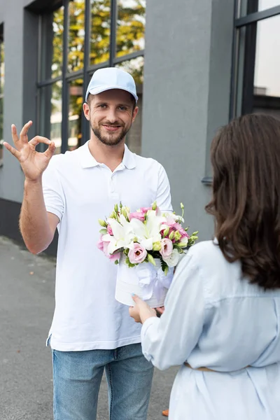 Messa a fuoco selettiva di felice consegna uomo in cap mostrando segno ok vicino donna con fiori — Foto stock