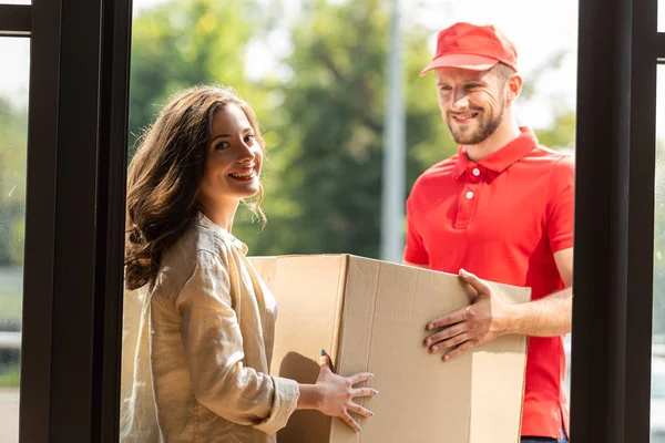 Cheerful delivery man in cap looking at camera and holding box near woman — Stock Photo