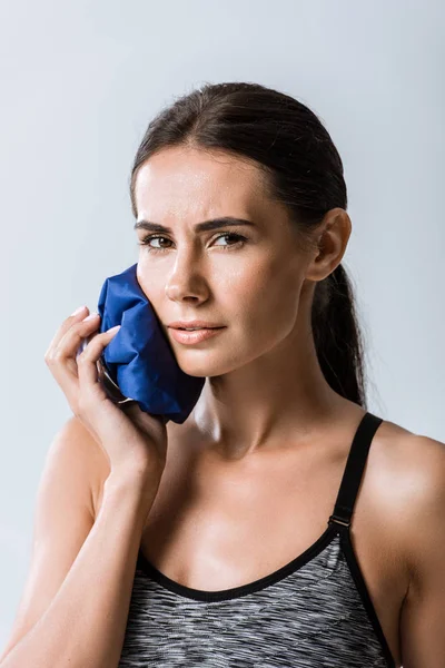 Attractive sportswoman using ice for cheek isolated on grey — Stock Photo