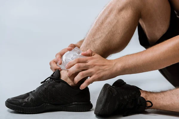 Частичный вид спортсмена в черных кроссовках, использующего лед для лодыжки на сером — стоковое фото