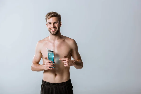 Vue de face d'un sportif barbu torse nu et souriant pointant du doigt une bouteille de sport isolée sur fond gris — Photo de stock