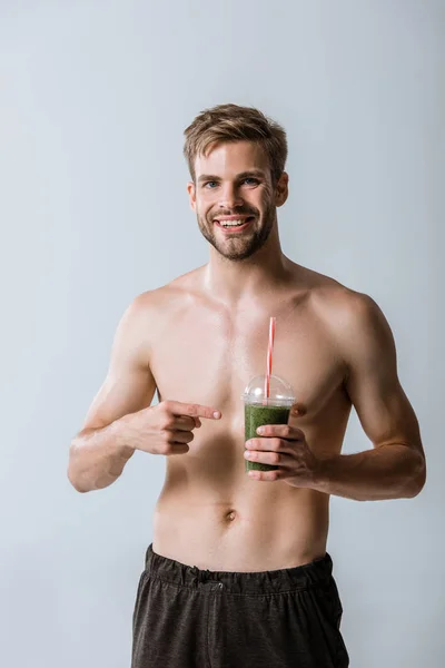 Front view of smiling shirtless sportsman pointing with finger at smoothie isolated on grey — Stock Photo