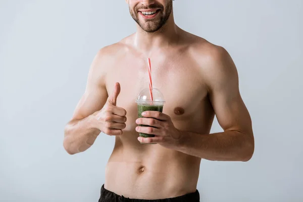 Partial view of smiling shirtless sportsman holding smoothie and showing thumb up isolated on grey — Stock Photo