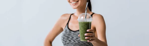 Panoramic shot of smiling sportswoman holding smoothie isolated on grey — Stock Photo