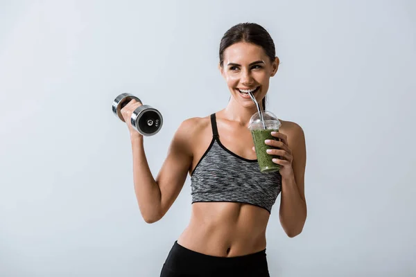 Smiling sportswoman holding dumbbell and drinking smoothie isolated on grey — Stock Photo