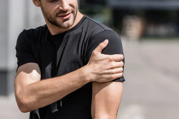 Vista cortada de desportista em t-shirt preta com dor no ombro na rua — Fotografia de Stock
