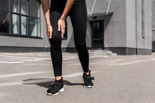 Vista ritagliata di sportiva in scarpe da ginnastica nere con dolore al ginocchio sulla strada — Foto stock
