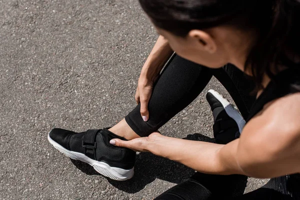 Vista ritagliata di sportiva in scarpe da ginnastica nere con dolore alla caviglia sulla strada — Foto stock