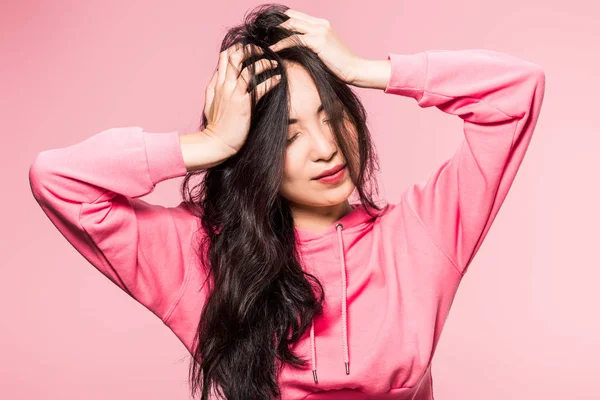 Attractive asian woman in pink pullover touching hair isolated on pink — Stock Photo