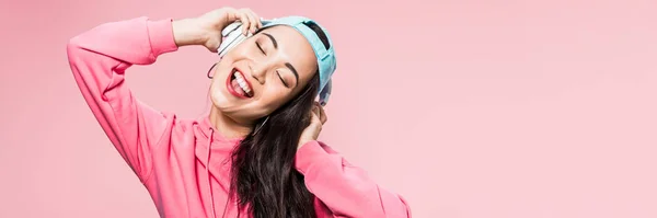 Panorâmica tiro de atraente asiático mulher no pulôver e boné ouvindo música e sorrindo isolado no rosa — Fotografia de Stock