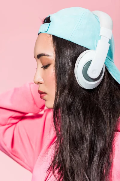 Atractiva mujer asiática en jersey y gorra escuchando música aislada en rosa - foto de stock