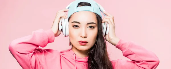 Plano panorámico de atractiva mujer asiática en jersey y gorra escuchando música aislada en rosa - foto de stock