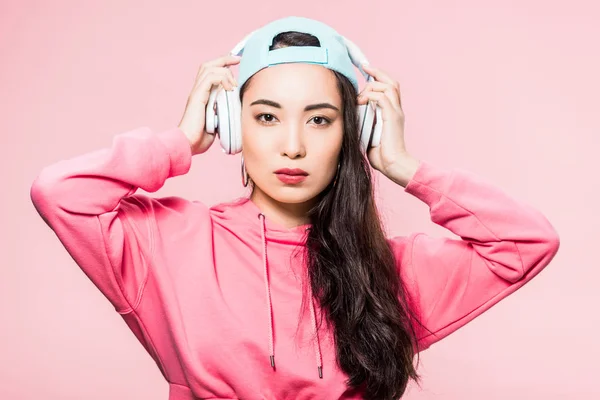 Atractiva mujer asiática en jersey y gorra escuchando música aislada en rosa - foto de stock