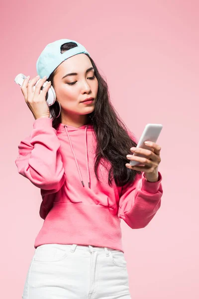 Atractiva mujer asiática en jersey y gorra escuchando música y sosteniendo teléfono inteligente aislado en rosa - foto de stock
