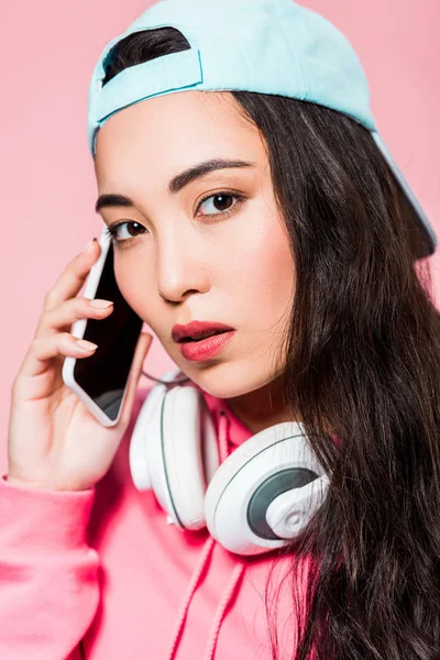 Attractive asian woman in pullover and cap with headphones talking on smartphone isolated on pink — Stock Photo