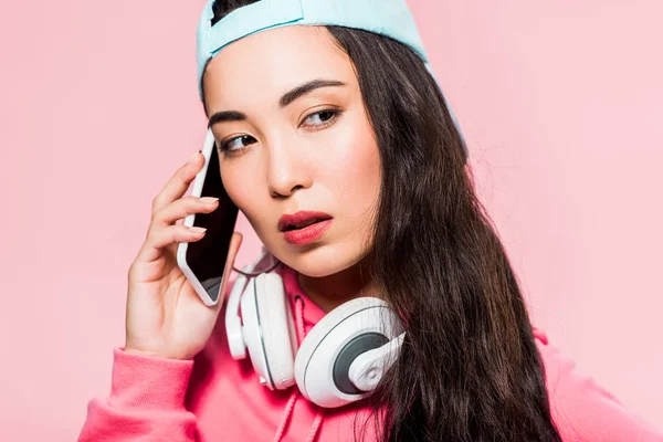 Atractiva mujer asiática en jersey y gorra con auriculares hablando en smartphone aislado en rosa - foto de stock