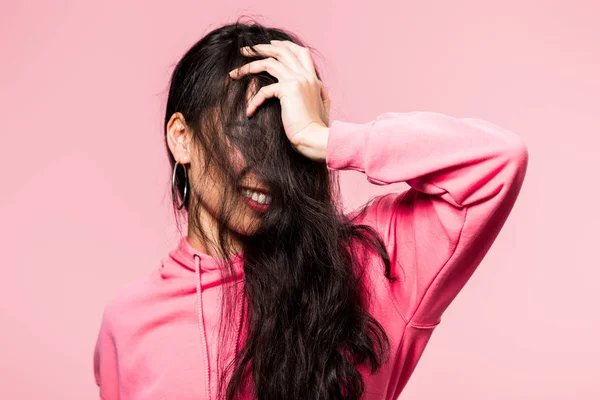 Atractiva mujer asiática en rosa jersey sonriendo y oscureciendo cara aislado en rosa - foto de stock