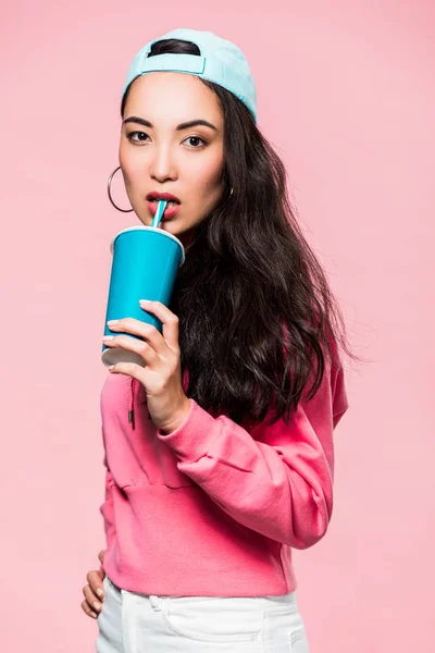 Atractiva mujer asiática en jersey y gorra bebiendo de taza de plástico aislado en rosa - foto de stock