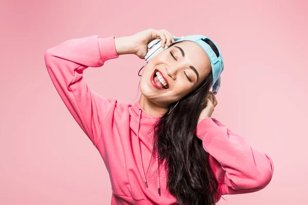 Atractiva mujer asiática en jersey y gorra escuchando música y sonriendo aislado en rosa - foto de stock