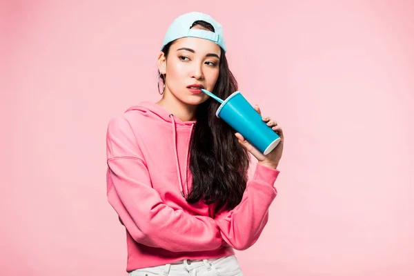 Pensive asian woman in pullover and cap drinking from plastic cup isolated on pink — Stock Photo