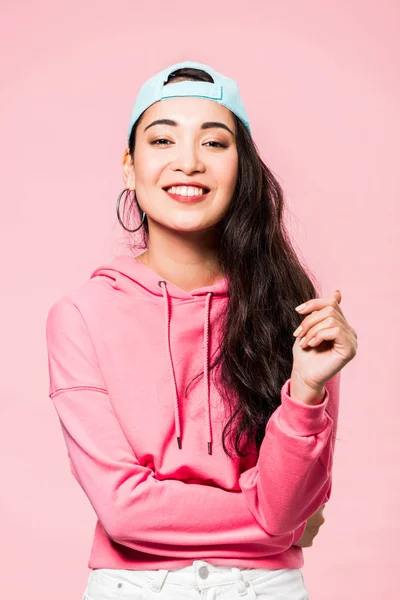 Atractiva mujer asiática en rosa jersey y gorra sonriente aislado en rosa - foto de stock