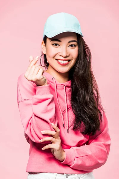 Attraente donna asiatica in pullover rosa e cappuccio sorridente e mostrando il simbolo del cuore isolato sul rosa — Foto stock