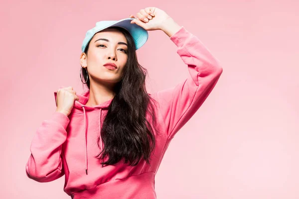 Atractiva y pensativa mujer asiática en suéter rosa y gorra mirando hacia otro lado aislado en rosa - foto de stock