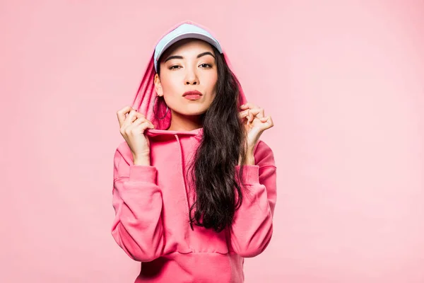 Attractive asian woman in pink pullover and cap looking at camera isolated on pink — Stock Photo