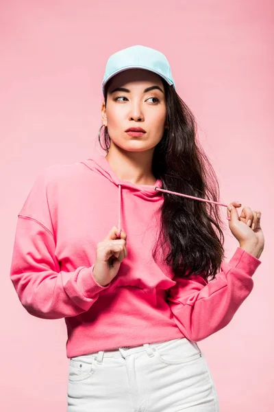 Atractiva y pensativa mujer asiática en suéter rosa y gorra mirando hacia otro lado aislado en rosa - foto de stock