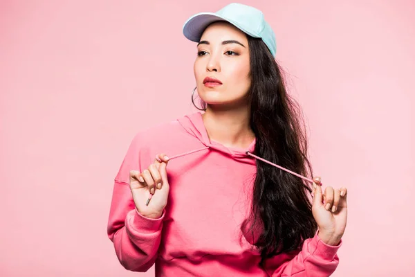 Atractivo asiático mujer en rosa jersey y gorra mirando lejos aislado en rosa - foto de stock