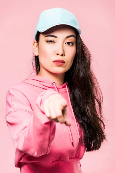 Foco seletivo de atraente mulher asiática em pulôver rosa e cap apontando com o dedo isolado em rosa — Fotografia de Stock