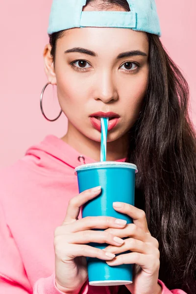 Attractive asian woman in pullover and cap drinking from plastic cup isolated on pink — Stock Photo