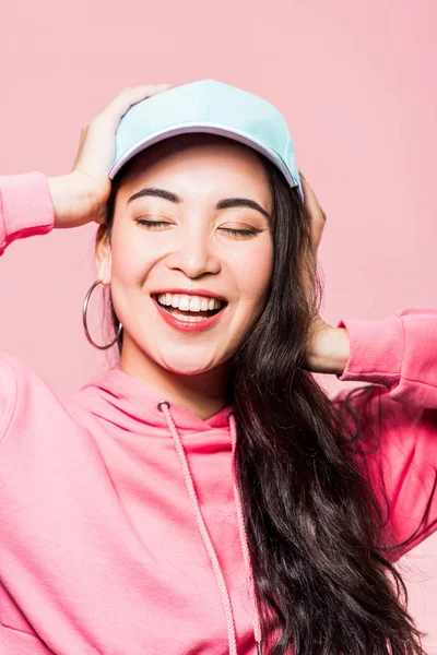 Atractiva mujer asiática con los ojos cerrados en suéter rosa y gorra sonriente aislado en rosa - foto de stock