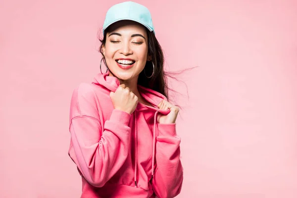 Atractiva mujer asiática con los ojos cerrados en suéter rosa y gorra sonriente aislado en rosa - foto de stock