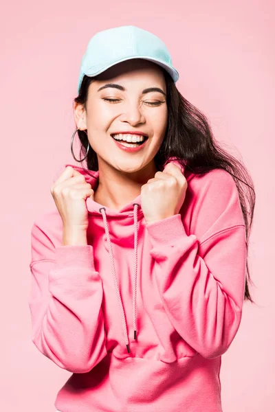 Attractive asian woman with closed eyes in pink pullover and cap smiling isolated on pink — Stock Photo