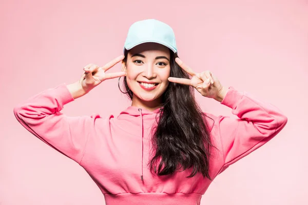 Attractive asian woman in pink pullover and cap smiling and showing peace sign isolated on pink — Stock Photo