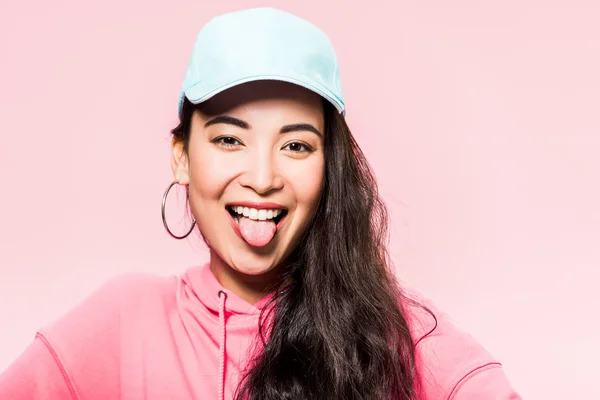 Attraktive asiatische Frau in rosa Pullover und Mütze abstehende Zunge isoliert auf rosa — Stockfoto