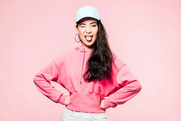 Attractive asian woman in pink pullover and cap winking and sticking out tongue isolated on pink — Stock Photo