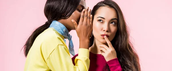 Foto panorámica de mujer afroamericana contando secreto a su amigo asiático aislado en rosa - foto de stock