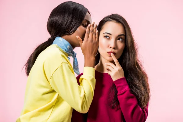 Africano americano mujer decir secreto a su asiático amigo aislado en rosa - foto de stock