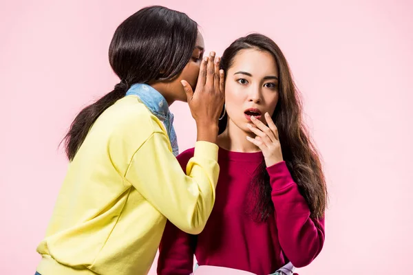 Africano mulher americana dizendo segredo para seu amigo asiático chocado isolado em rosa — Fotografia de Stock